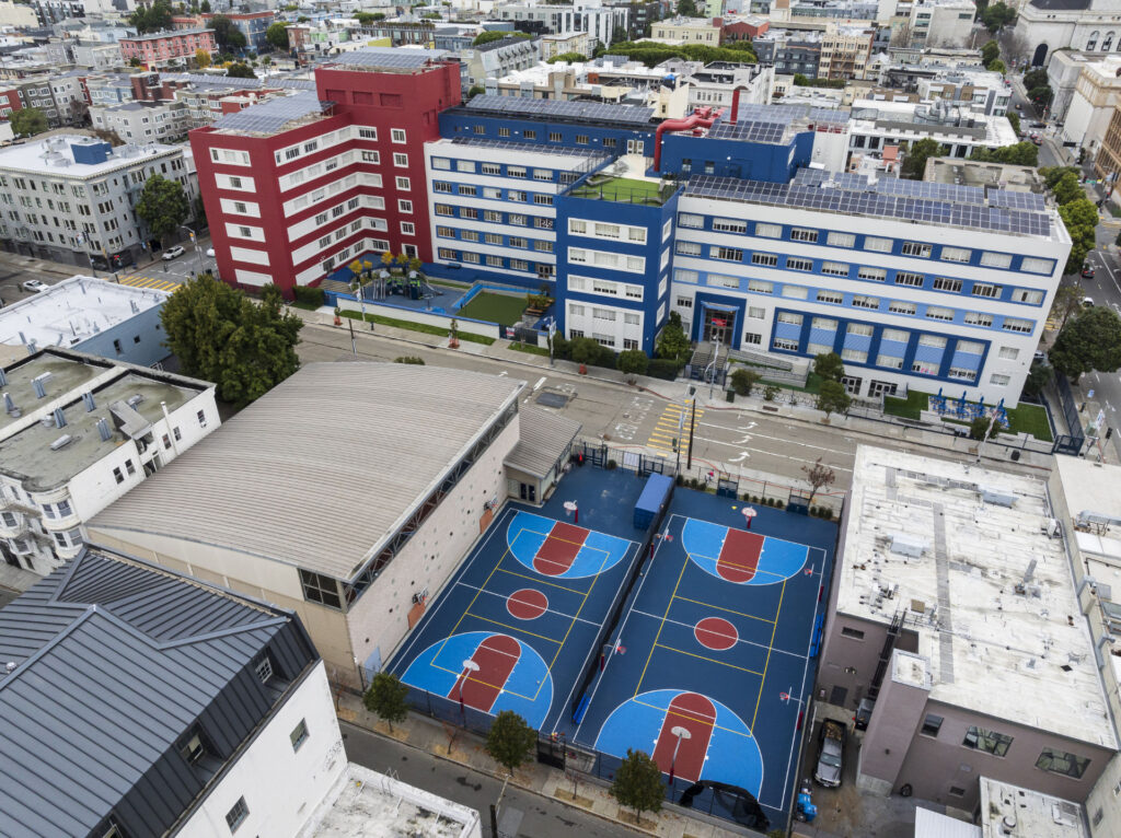 Aerial view of two full sized basketball courts at International School of San Francisco with various city buildings in the background. The basketball courts have been resurfaced using Laykold acrylic Dark Blue, Light Blue and Red. Yellow volleyball lines have been painted inside both basketball courts.
